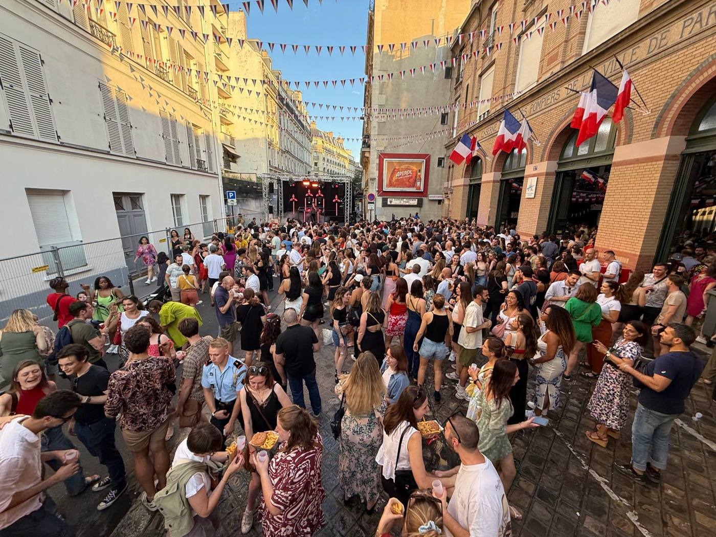 Foule dansant dans la rue sous les fanions bleu-blanc-rouge, scène au fond, façade de la caserne à droite.
