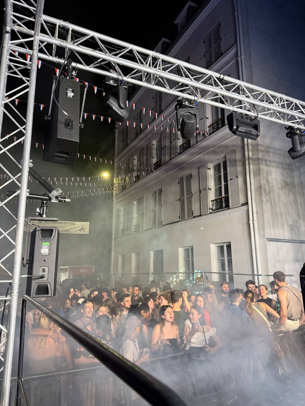 De nuit : enceintes et projecteurs asservis suspendus à la structure, fumée et foule derrière les barrières, banderole Bal des pompiers de Paris.