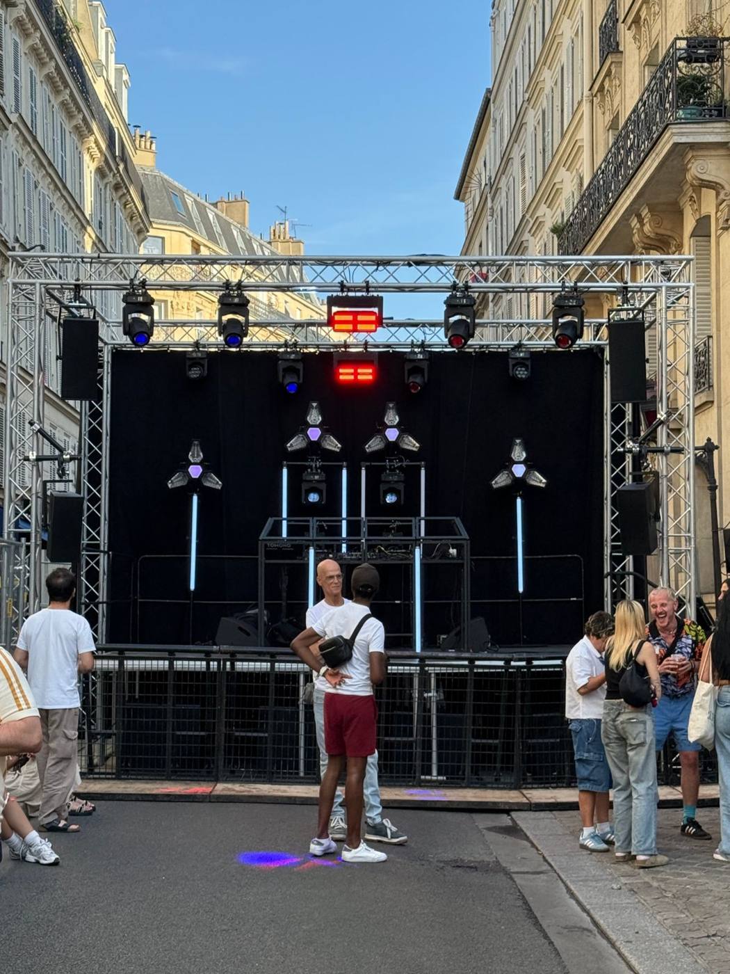 Scène en rue avant l'ouverture : structure en aluminium, projecteurs asservis, barres lumineuses et enceintes suspendues sur fond noir.