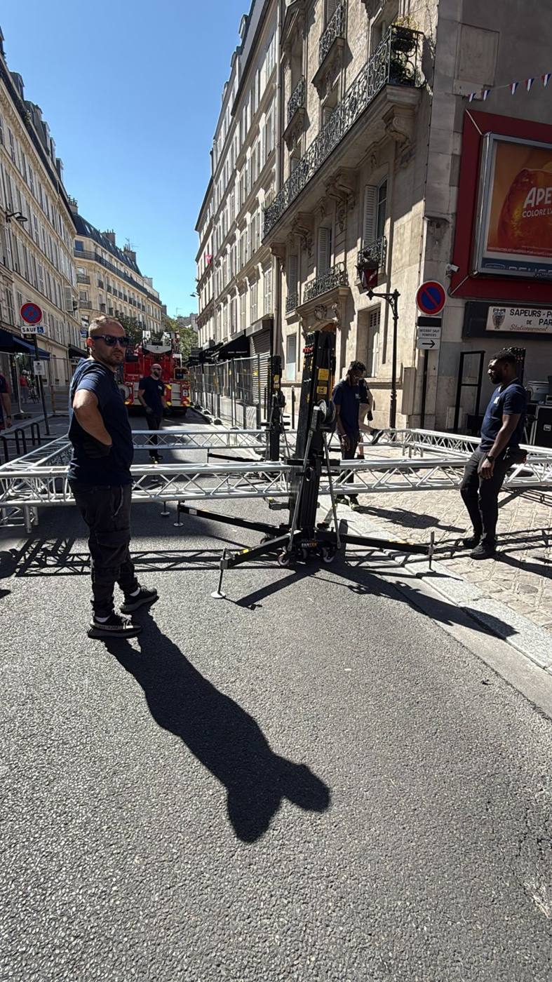 Montage : équipe autour d'un élévateur à structure, poutrelles au sol dans la rue, échelle de pompiers au fond.