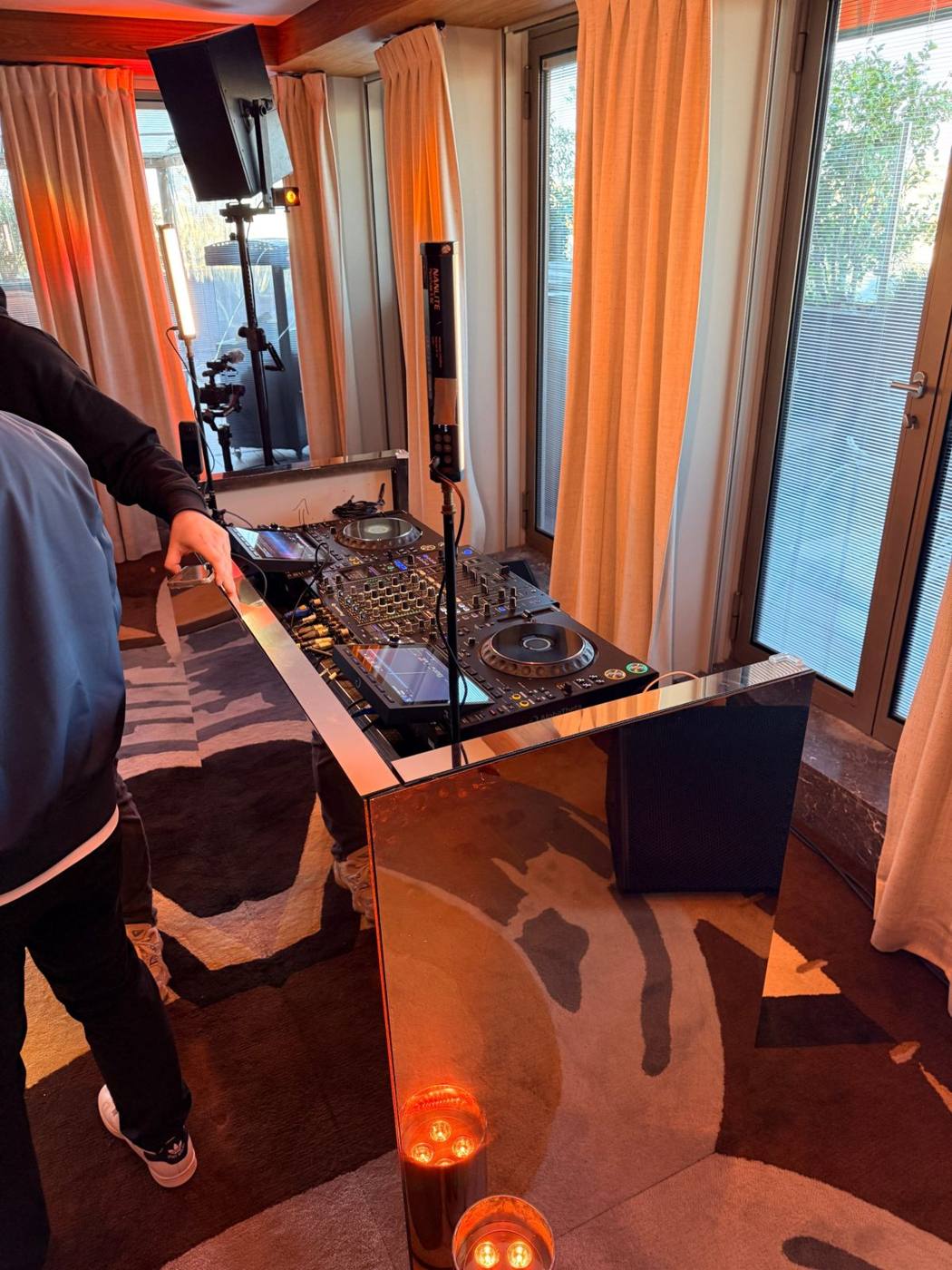Table de DJ en miroir cuivré, platines et mixeur, enceinte sur pied et éclairage tube, rideaux orangés.