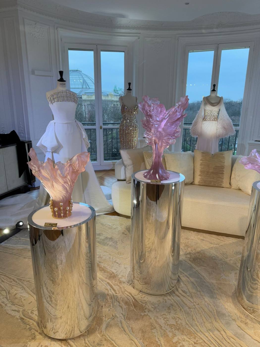 Salon d'angle avec fenêtres sur Paris : mannequins en robes de couture, sculptures en cristal rose sur piédestaux miroir.