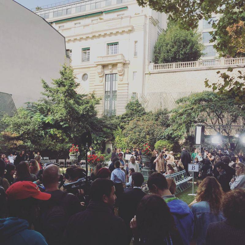 Jardin d'un hôtel particulier avec fauteuils pliants et public massé, projecteurs et caméras au fond.