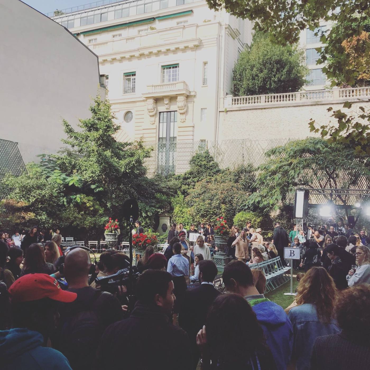 Jardin d'un hôtel particulier avec fauteuils pliants et public massé, projecteurs et caméras au fond.