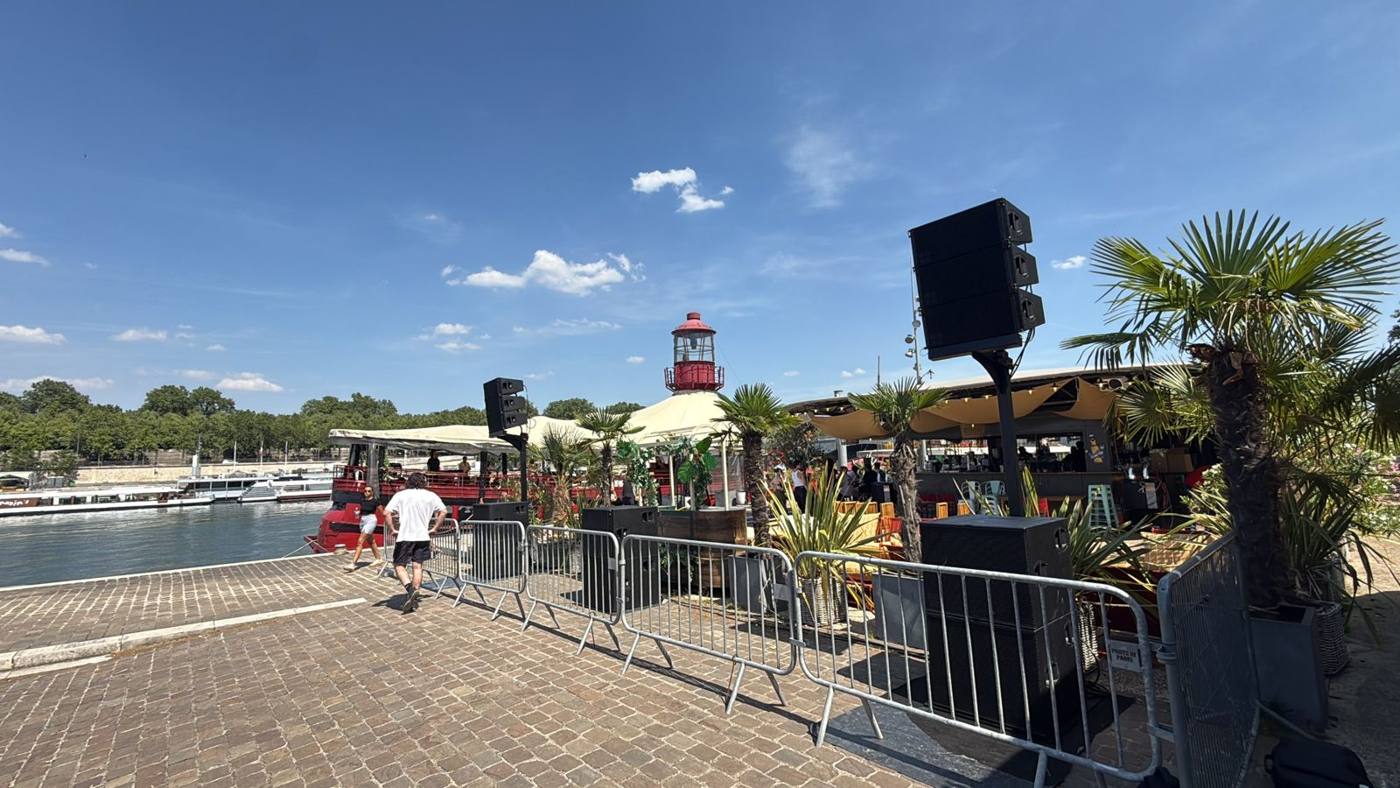 Vue du quai : enceintes sur pied derrière des barrières, palmiers et péniche-bar.