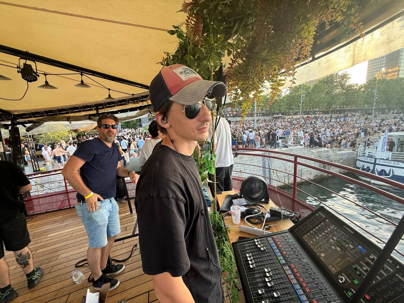 Ingénieur du son à la console Yamaha sur le pont d'une péniche, foule sur le quai en contrebas.