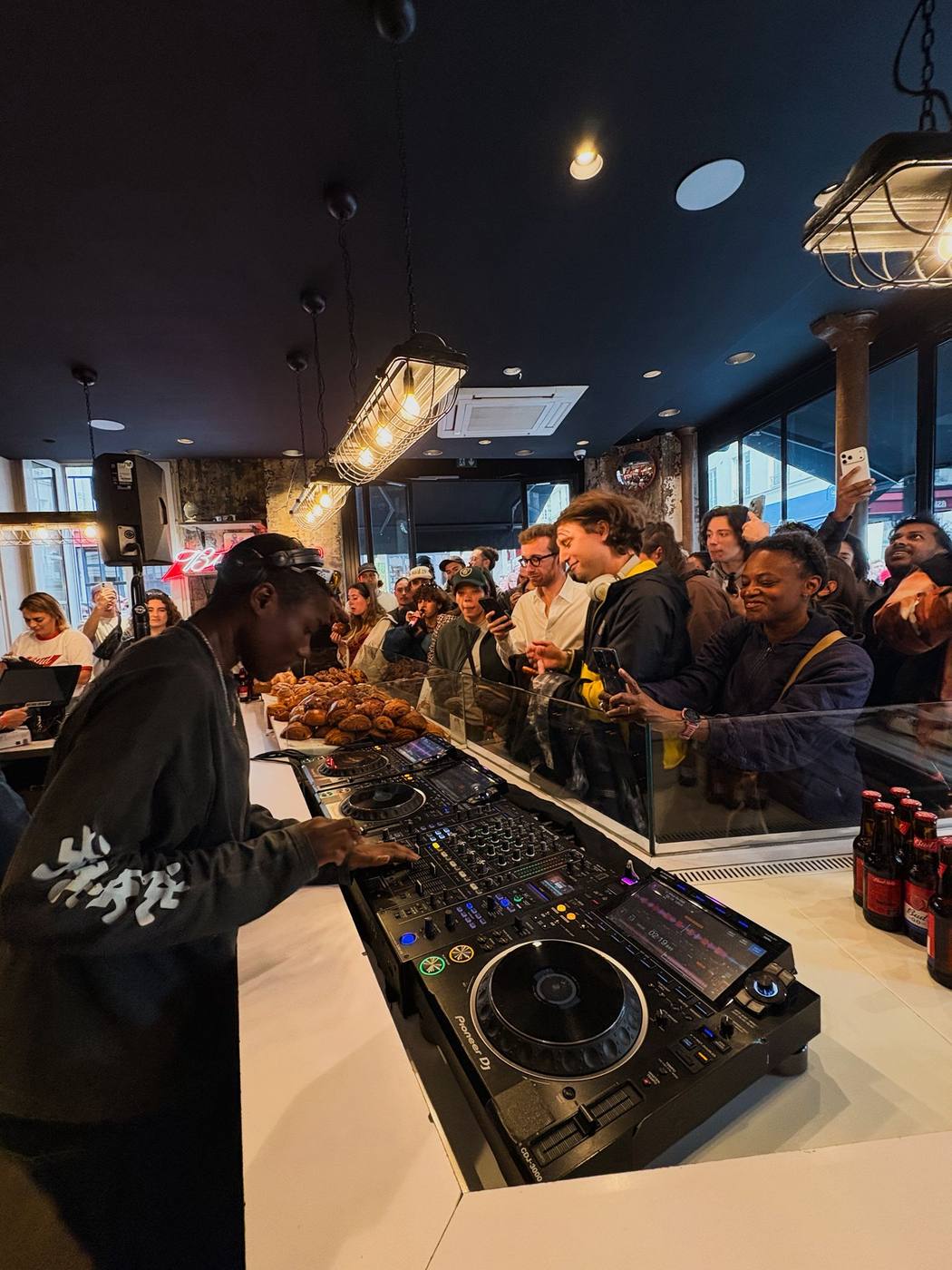 DJ en sweat sombre aux platines Pioneer sur le comptoir blanc d'une boulangerie, public massé derrière la vitrine, enceinte sur pied et bouteilles Bud 0.0.