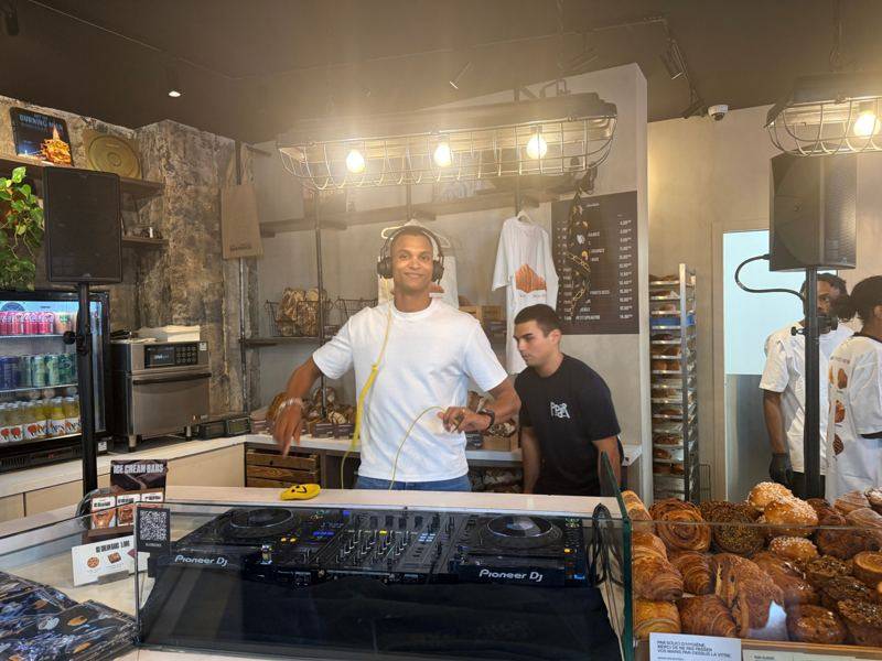 DJ casque sur les oreilles derrière des platines Pioneer DJ, dans une boulangerie, enceintes sur pieds de chaque côté.