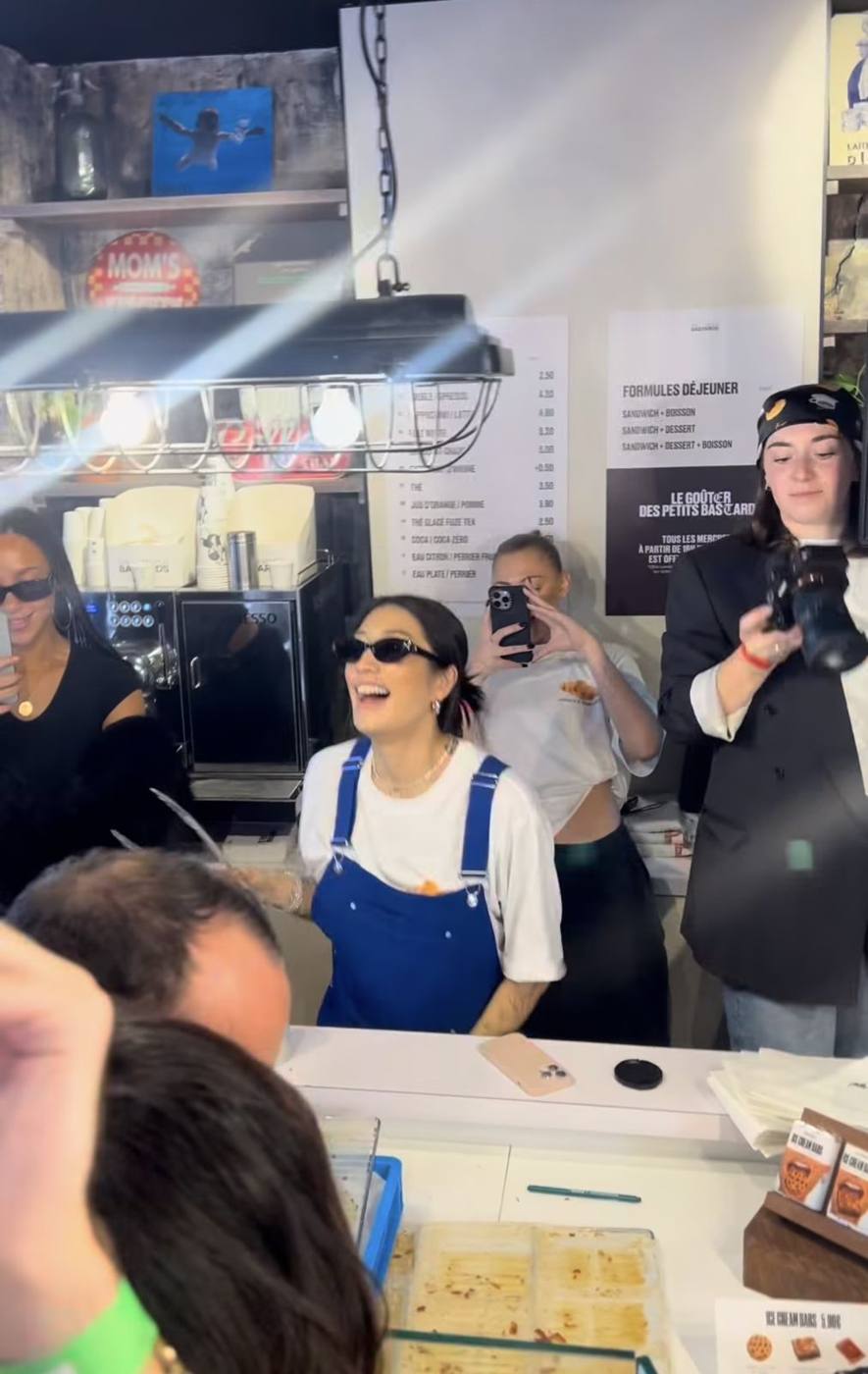 Peggy Gou souriante derrière le comptoir de la boulangerie, entourée de personnes qui filment.