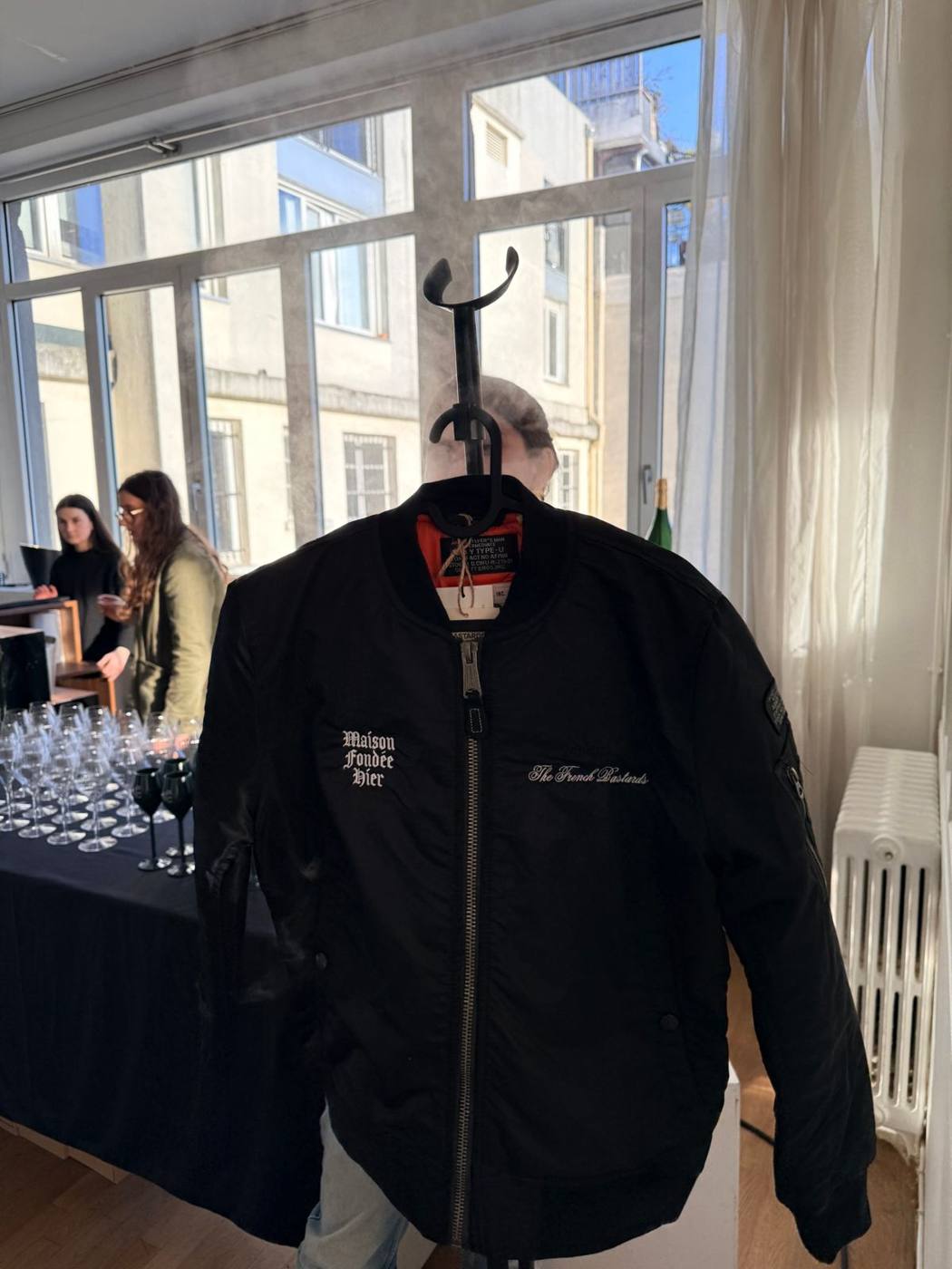 Blouson noir brodé « Maison Fondée Hier » et « The French Bastards » sur un cintre, verres en arrière-plan.
