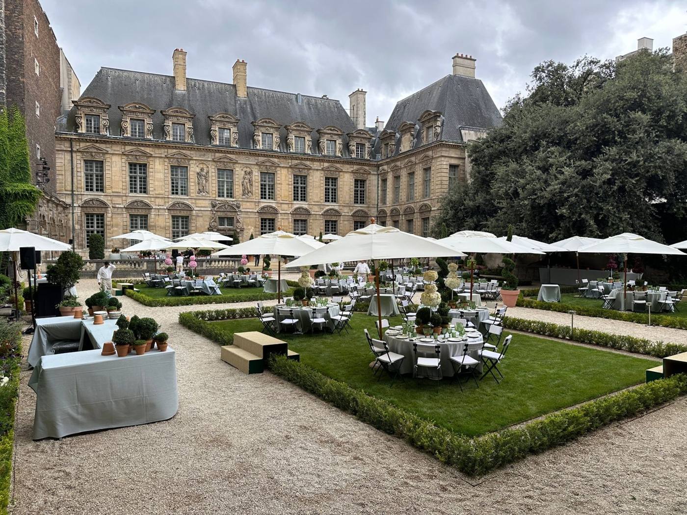 Jardin de l'hôtel particulier avec tables rondes, parasols blancs et buis taillés.