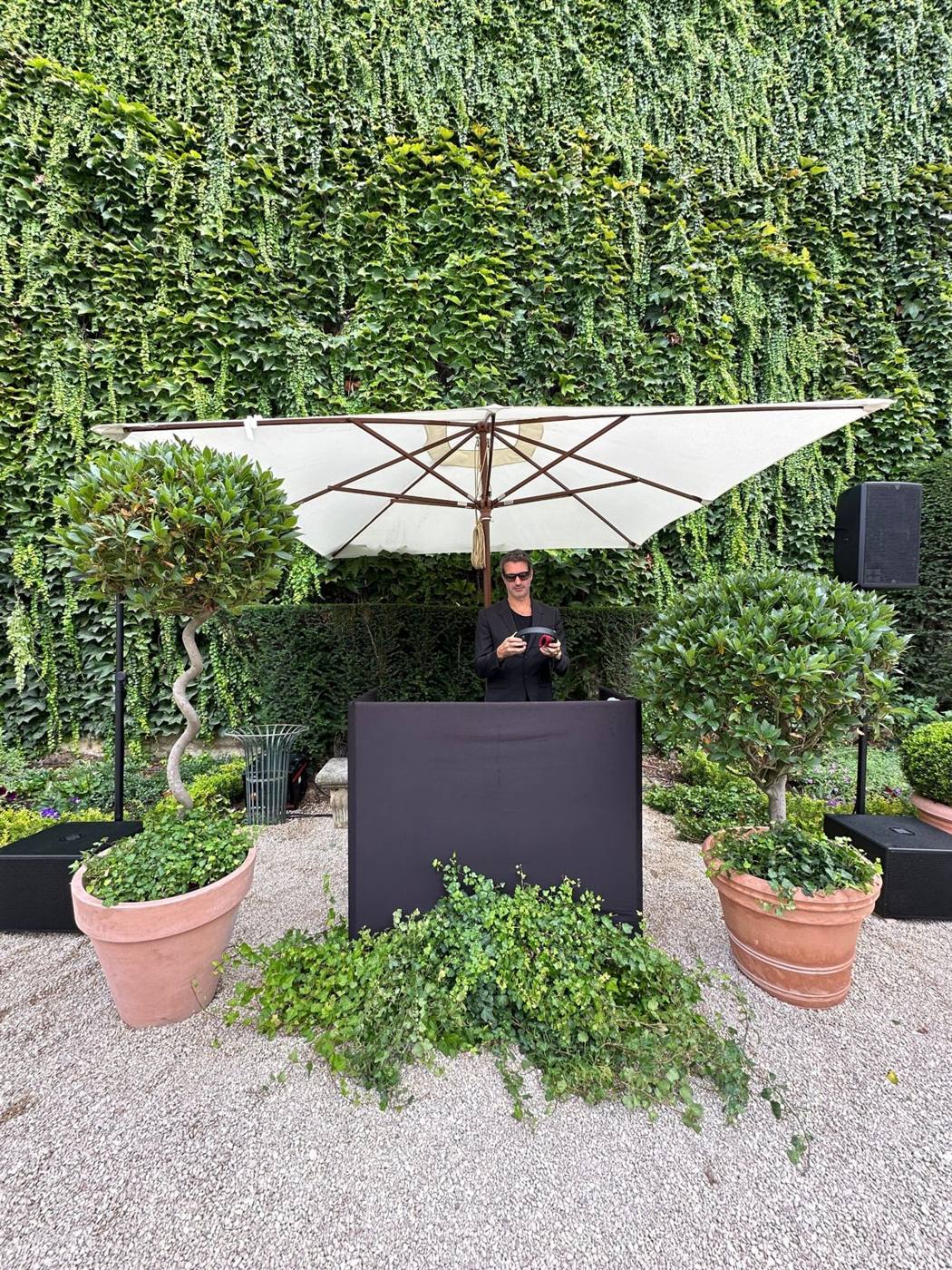 DJ derrière son pupitre sous un parasol, devant un mur de lierre, enceintes de chaque côté.