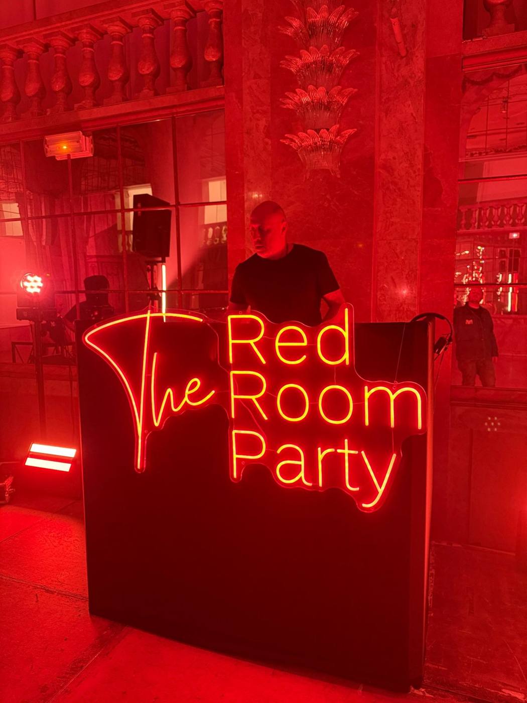 DJ derrière un pupitre noir portant le néon « The Red Room Party », colonnes et balustrades éclairées en rouge.