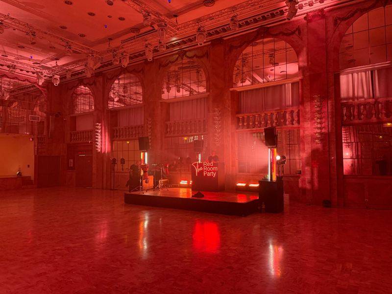 Grande salle éclairée en rouge : estrade, percussions, DJ et lyres suspendues sur structure.