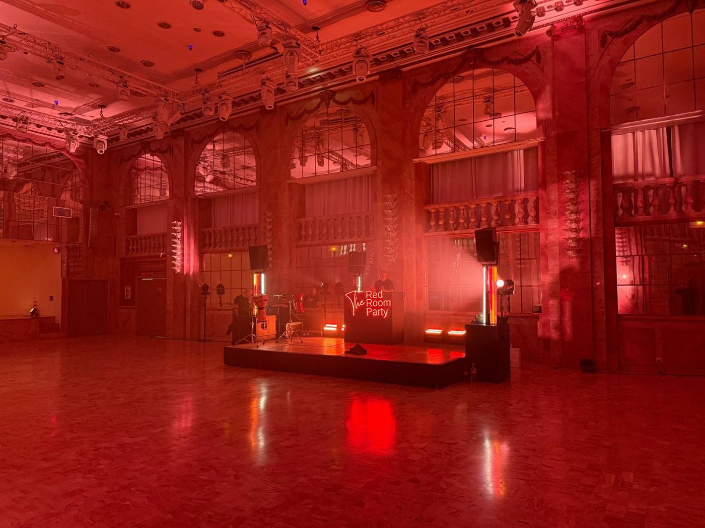 Grande salle éclairée en rouge : estrade, percussions, DJ et lyres suspendues sur structure.