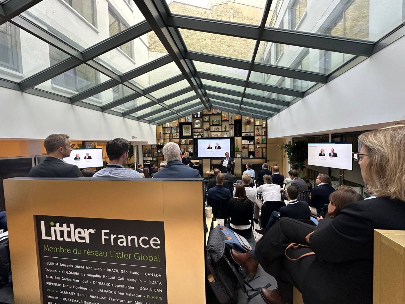 Intervenant devant un public assis sous la verrière, écrans de présentation dans la salle.
