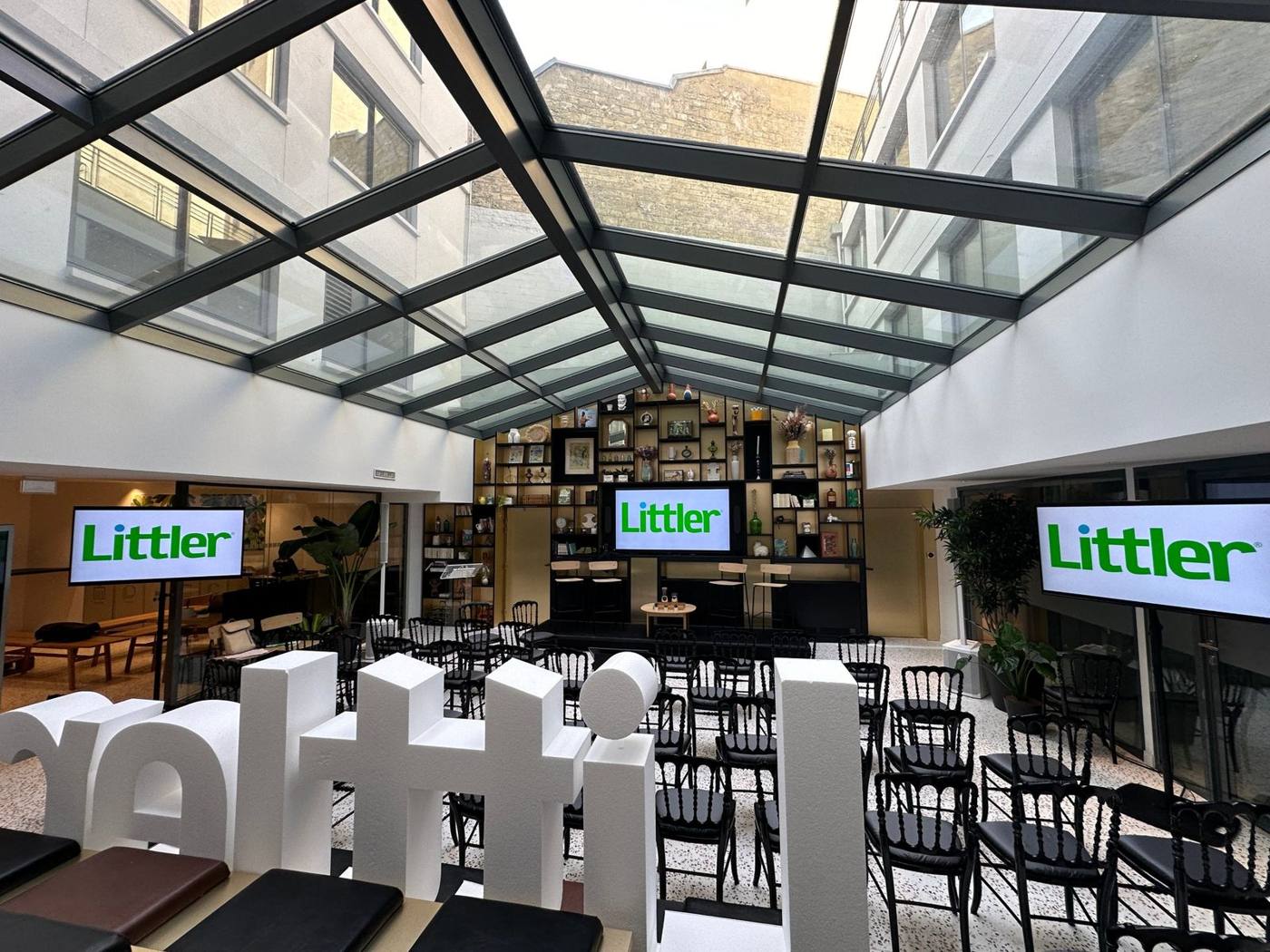 Salle de conférence sous une verrière : trois écrans au logo Littler, rangées de chaises noires, scène au fond.