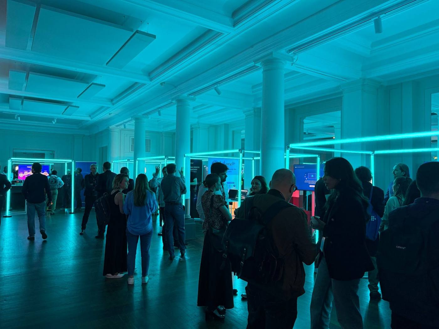 Foyer éclairé en cyan avec cadres lumineux et stands, visiteurs en discussion.