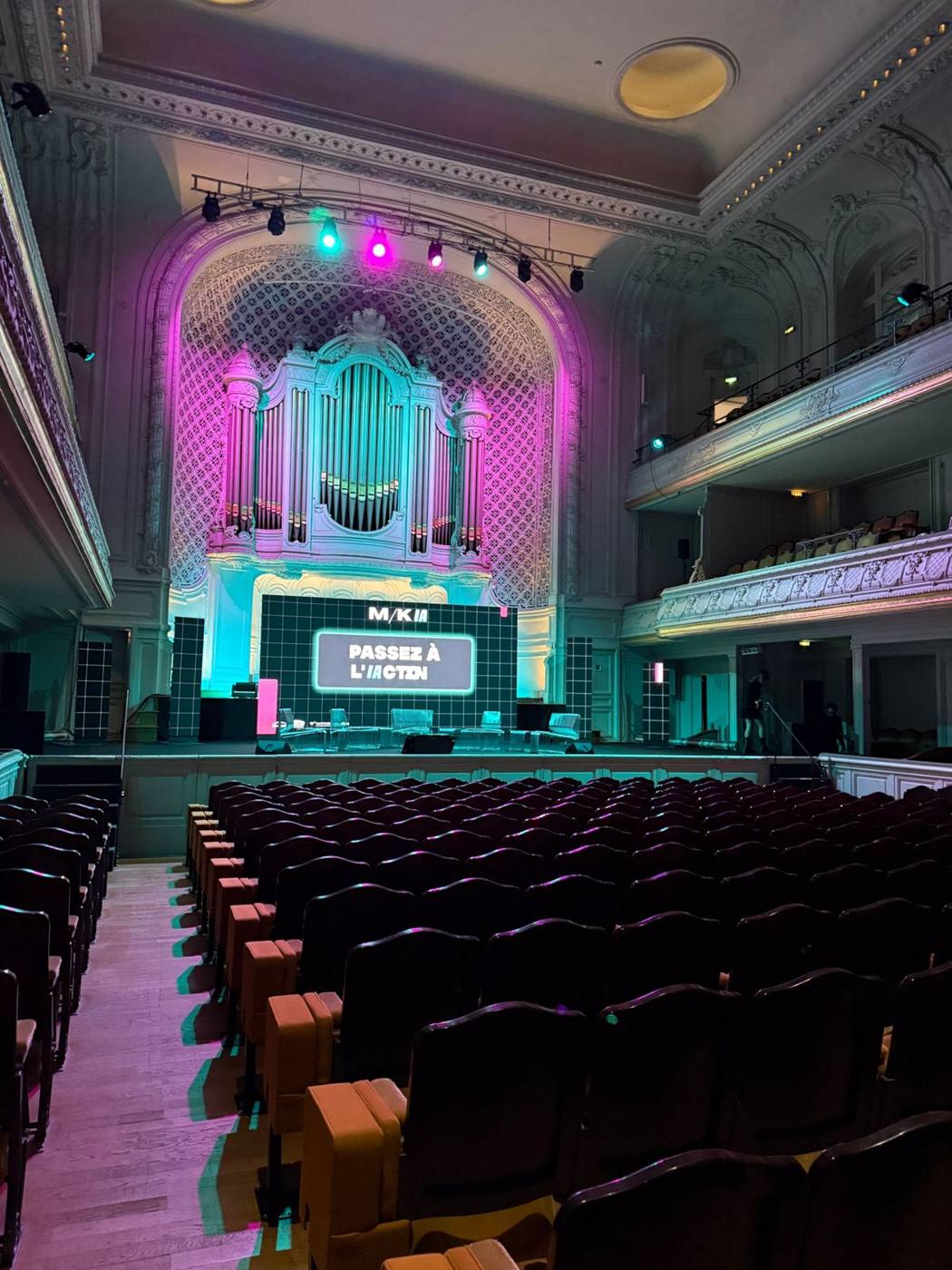 Salle vide avant l'ouverture : scène avec mur LED « M/K IA », orgue éclairé en rose et cyan, rangées de fauteuils.