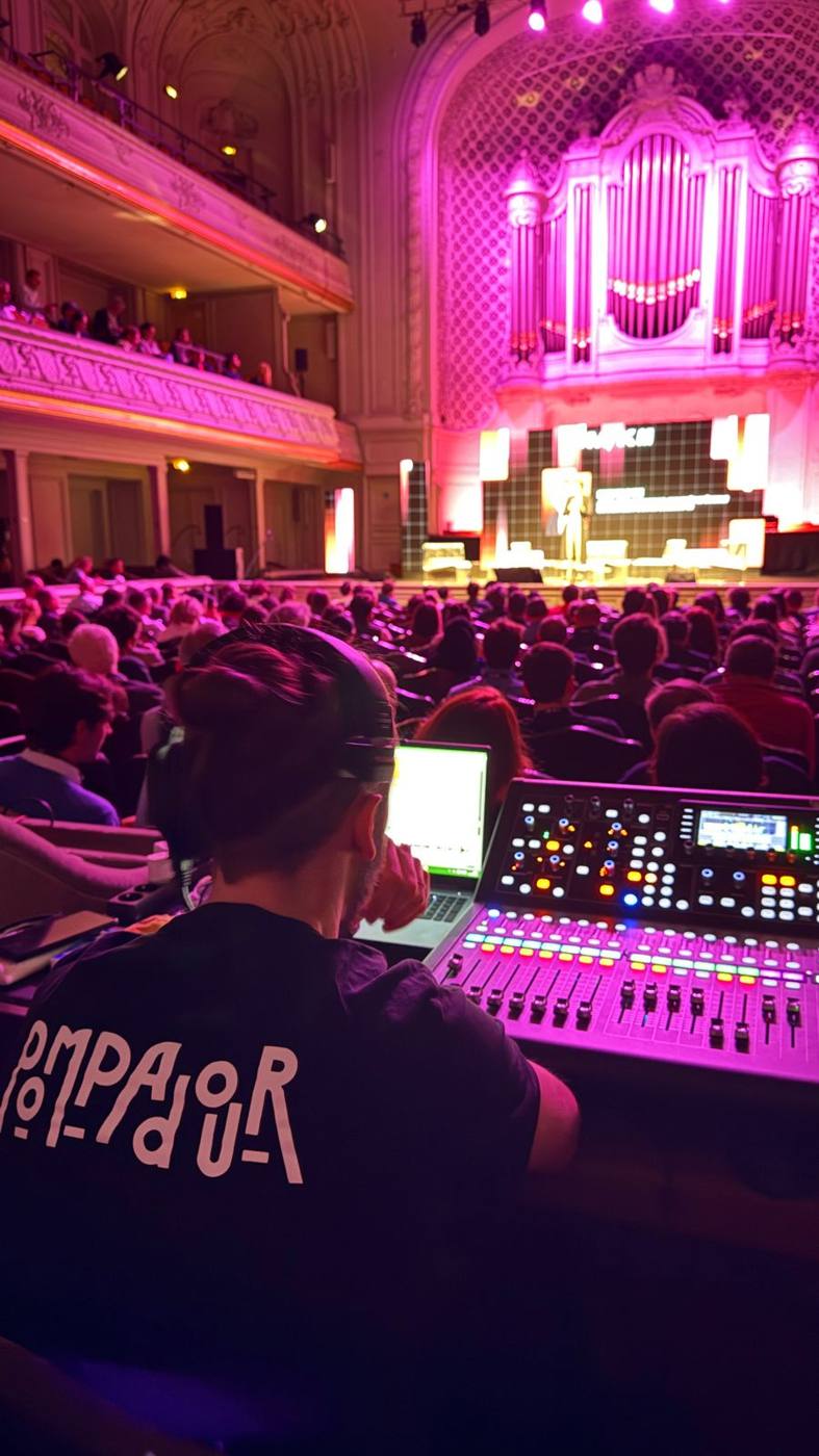 Technicien en t-shirt Pompadour à la console de mixage, salle de concert Art nouveau éclairée en rose et public assis.