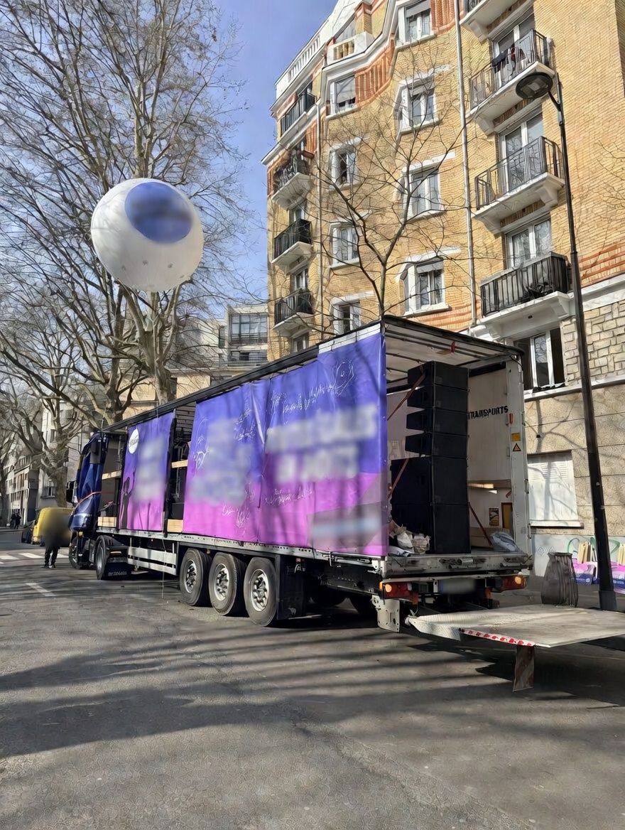 Semi-remorque aménagé en scène sonorisée, bâches violettes et ballon blanc et bleu au-dessus, rue parisienne.