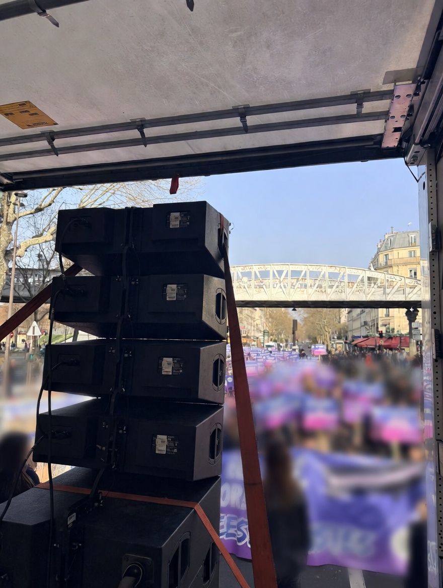 Colonne d'enceintes line array et caissons de grave sur le plateau, cortège et pont du métro aérien en arrière-plan.
