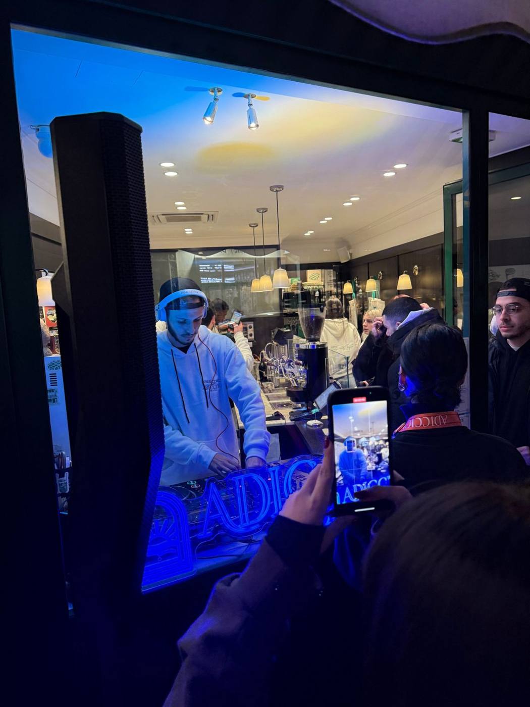 DJ in a white sweatshirt behind a café window, blue "ADICCION" neon in the foreground, crowd filming with phones.