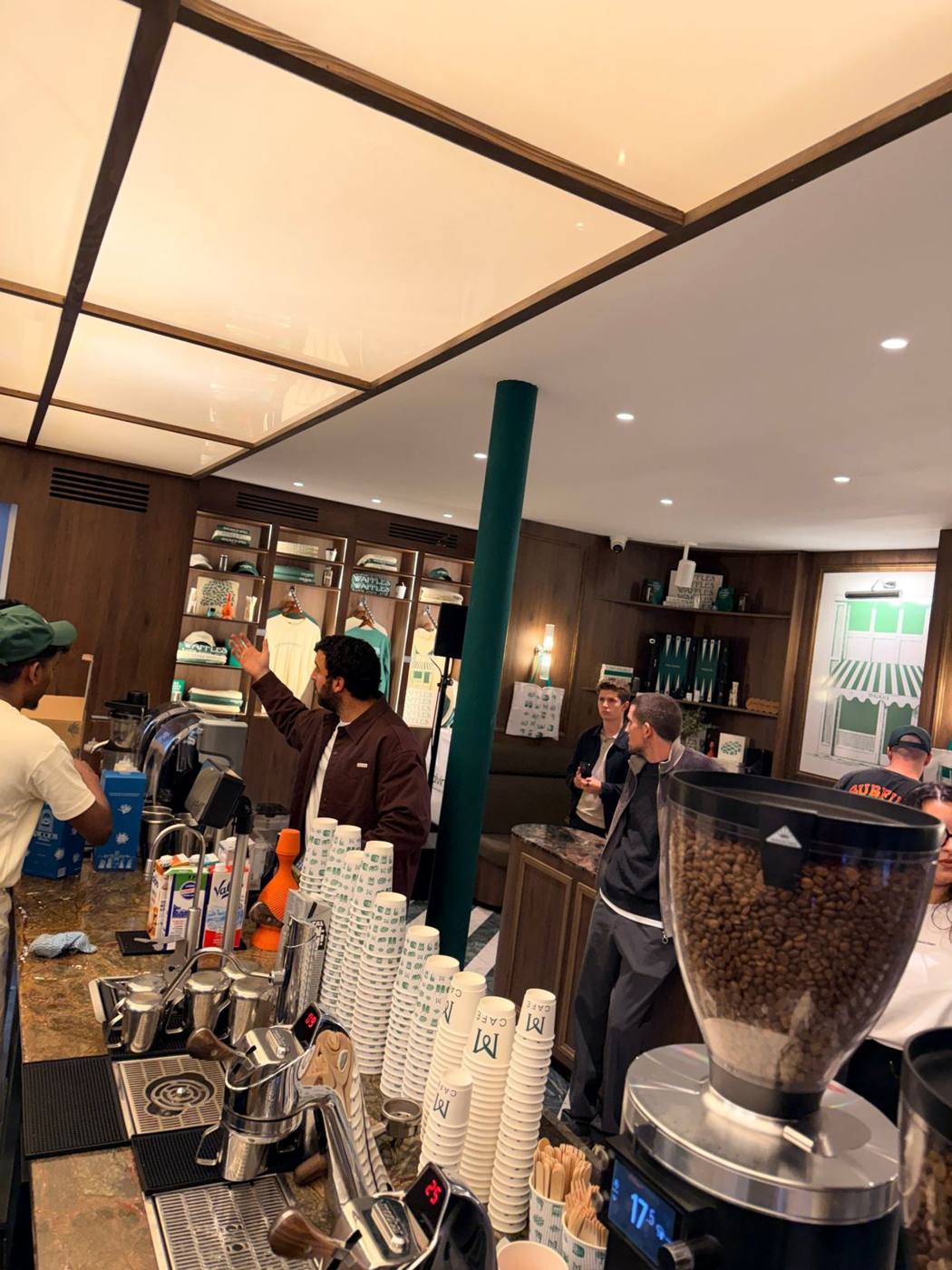 Comptoir du café : machine à espresso, gobelets empilés et moulin à café, étagères en bois en arrière-plan.