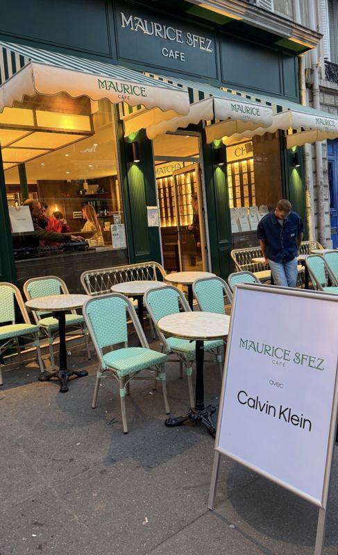 Façade du café Maurice Sfez, auvents rayés, terrasse et panneau « avec Calvin Klein ». Une régie DJ est visible en vitrine.