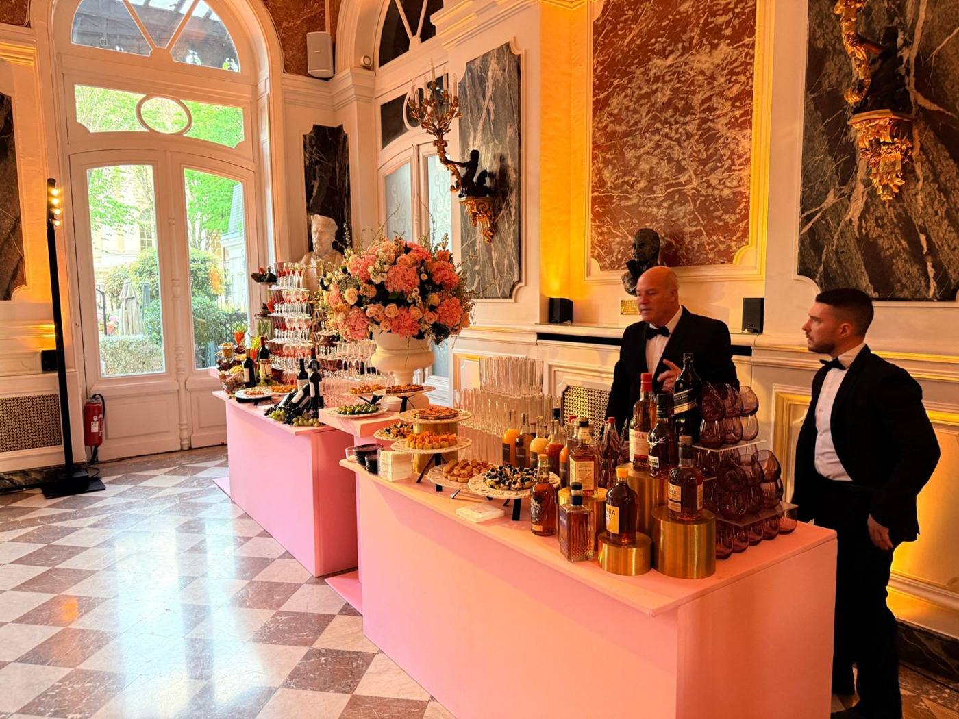Buffets sur tables roses, bouquet central, serveurs en nœud papillon, salon de marbre.