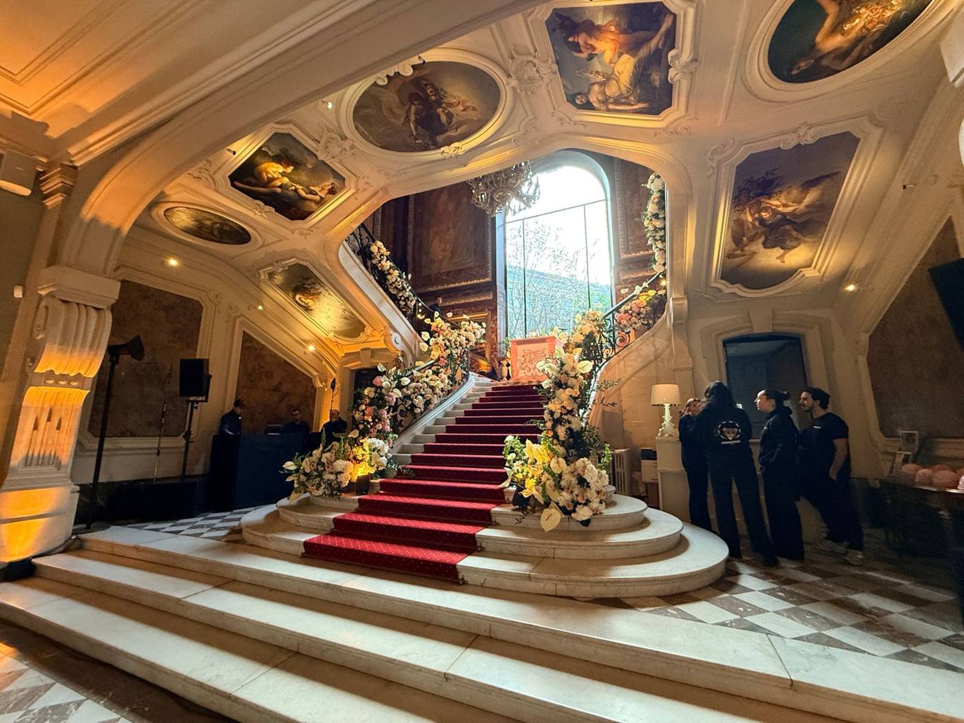 Hall et escalier d'honneur fleuri, plafond peint, équipe en noir à droite.
