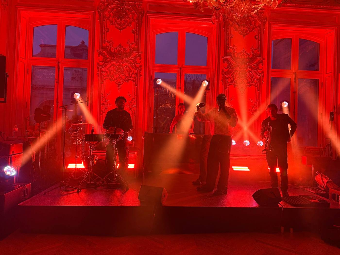 Scène éclairée en rouge : percussionniste, chanteur et trompettiste, faisceaux de projecteurs.