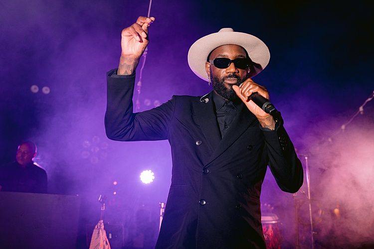 Chanteur en costume noir et chapeau clair au micro, éclairé en violet dans la fumée.