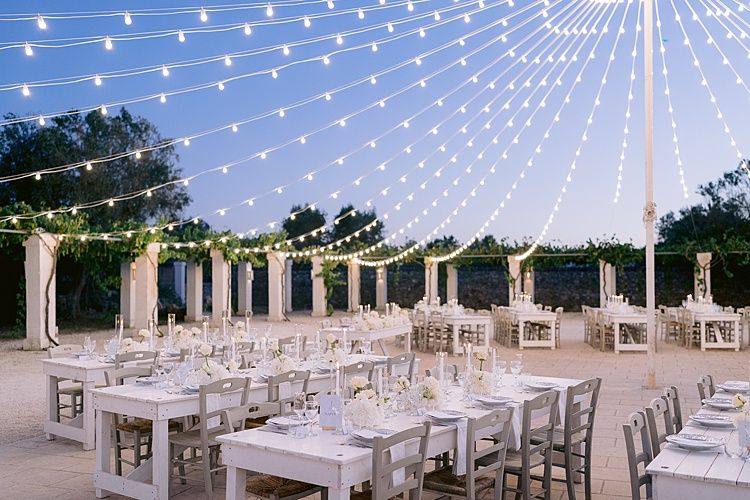 Tables dressées en blanc sous les guirlandes lumineuses, au crépuscule.