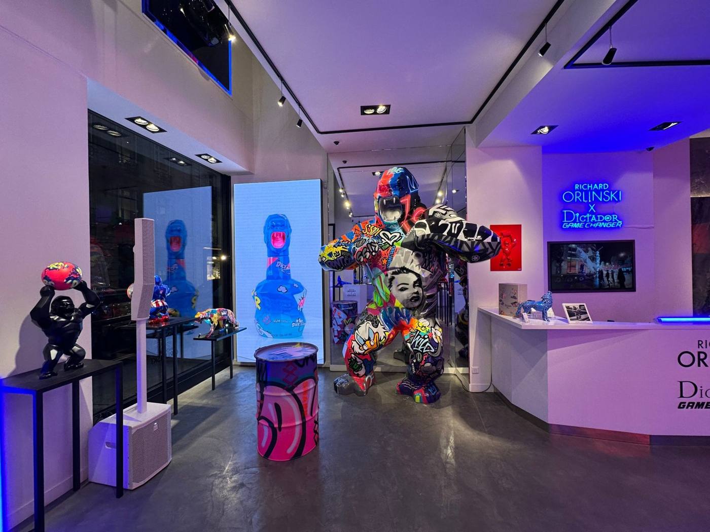 Galerie Orlinski : gorille sculpté multicolore, enseigne néon bleue, écran vertical, colonne de sonorisation et lumière violette.
