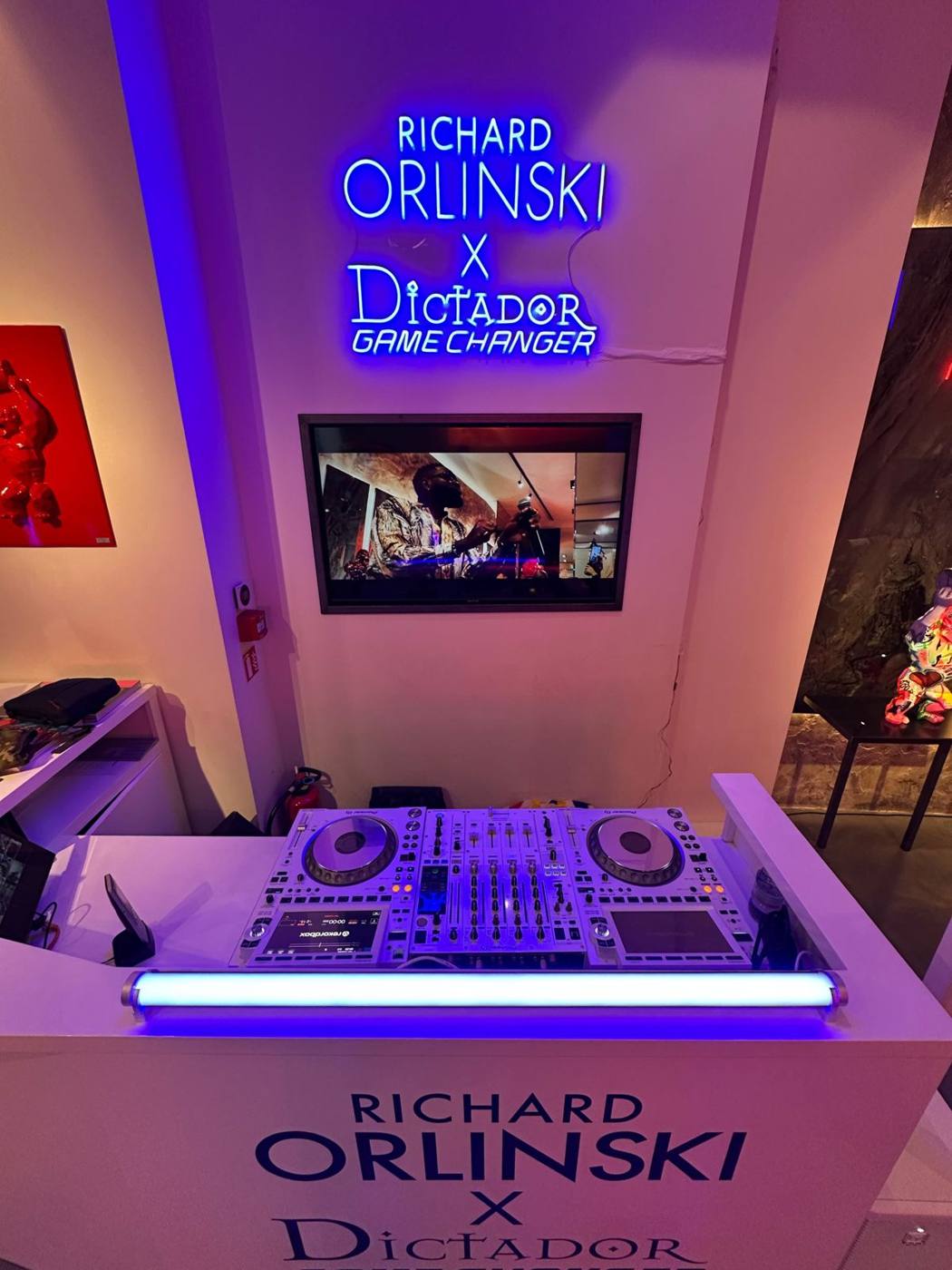 Pupitre de DJ avec contrôleur Pioneer et tube lumineux, sous l'enseigne néon Orlinski × Dictador et un écran vidéo.