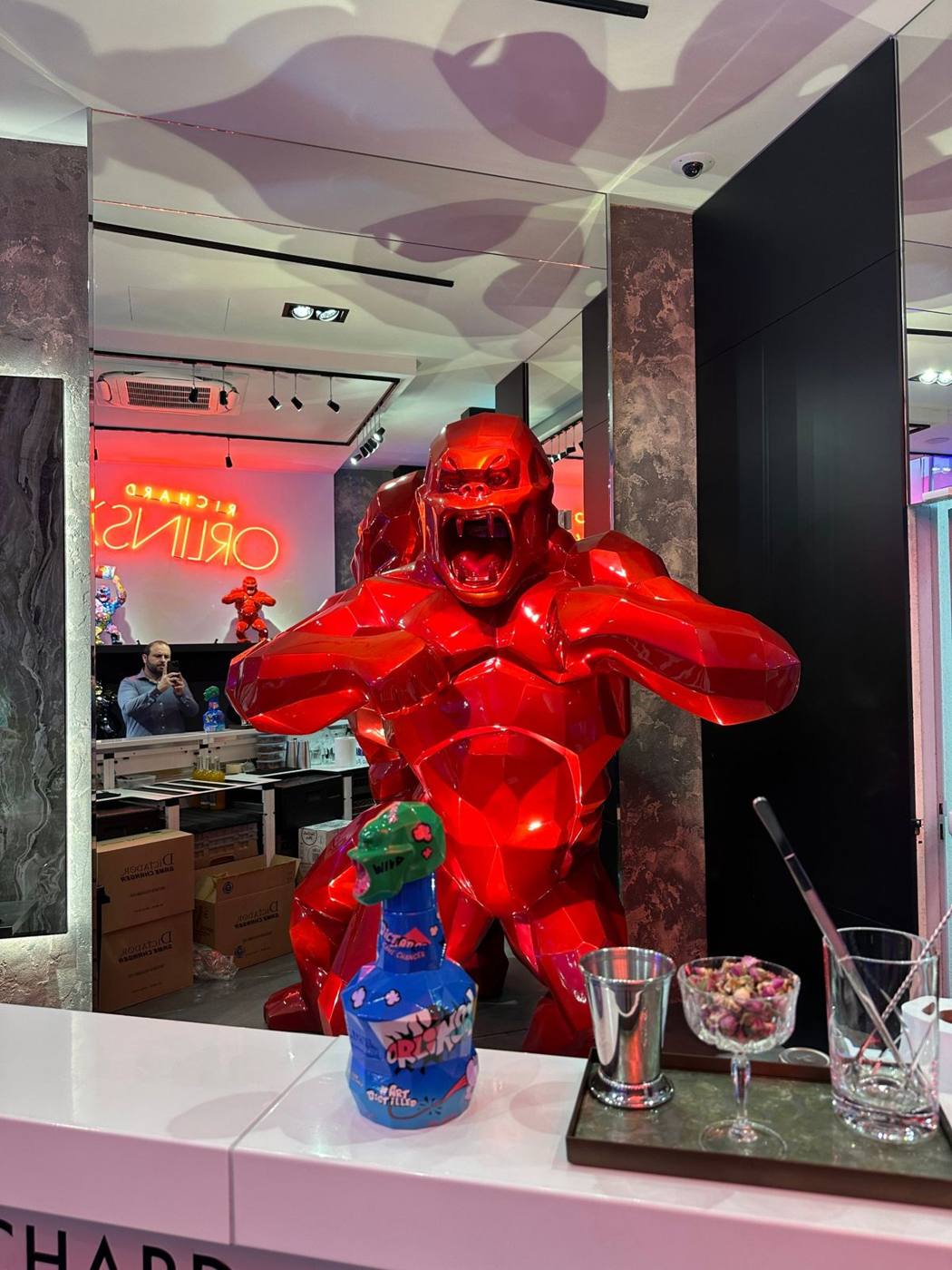 Gorille rouge brillant derrière le bar, bouteille Dictador au premier plan, enseigne néon rouge au fond.