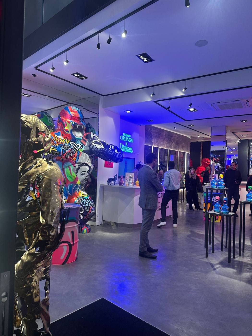 Invités dans la galerie : gorille multicolore à gauche, enseigne néon au fond, bouteilles exposées sur des guéridons.