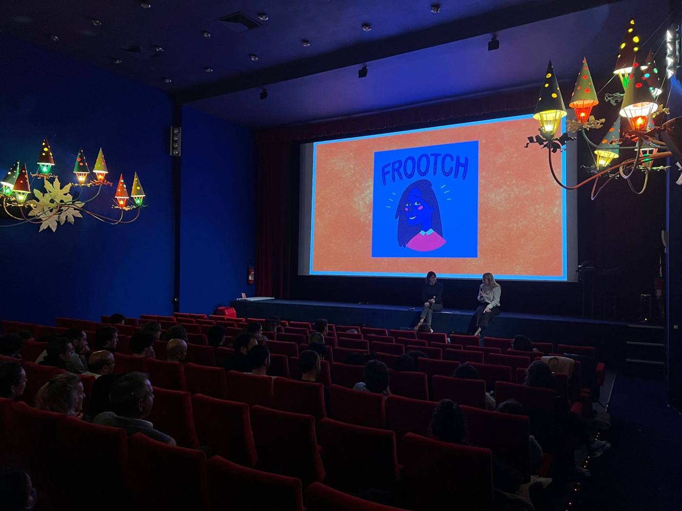 Salle de cinéma aux fauteuils rouges : deux intervenantes sur scène devant un écran orange.