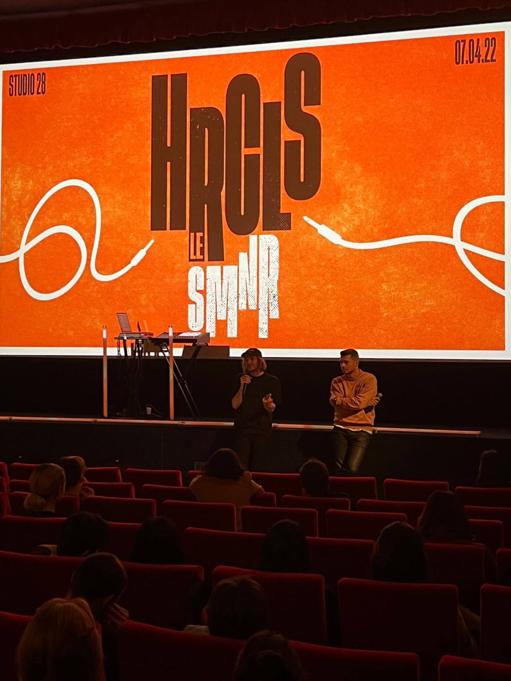 Écran de cinéma affichant l'identité du séminaire HRCLS, deux intervenants sur scène.