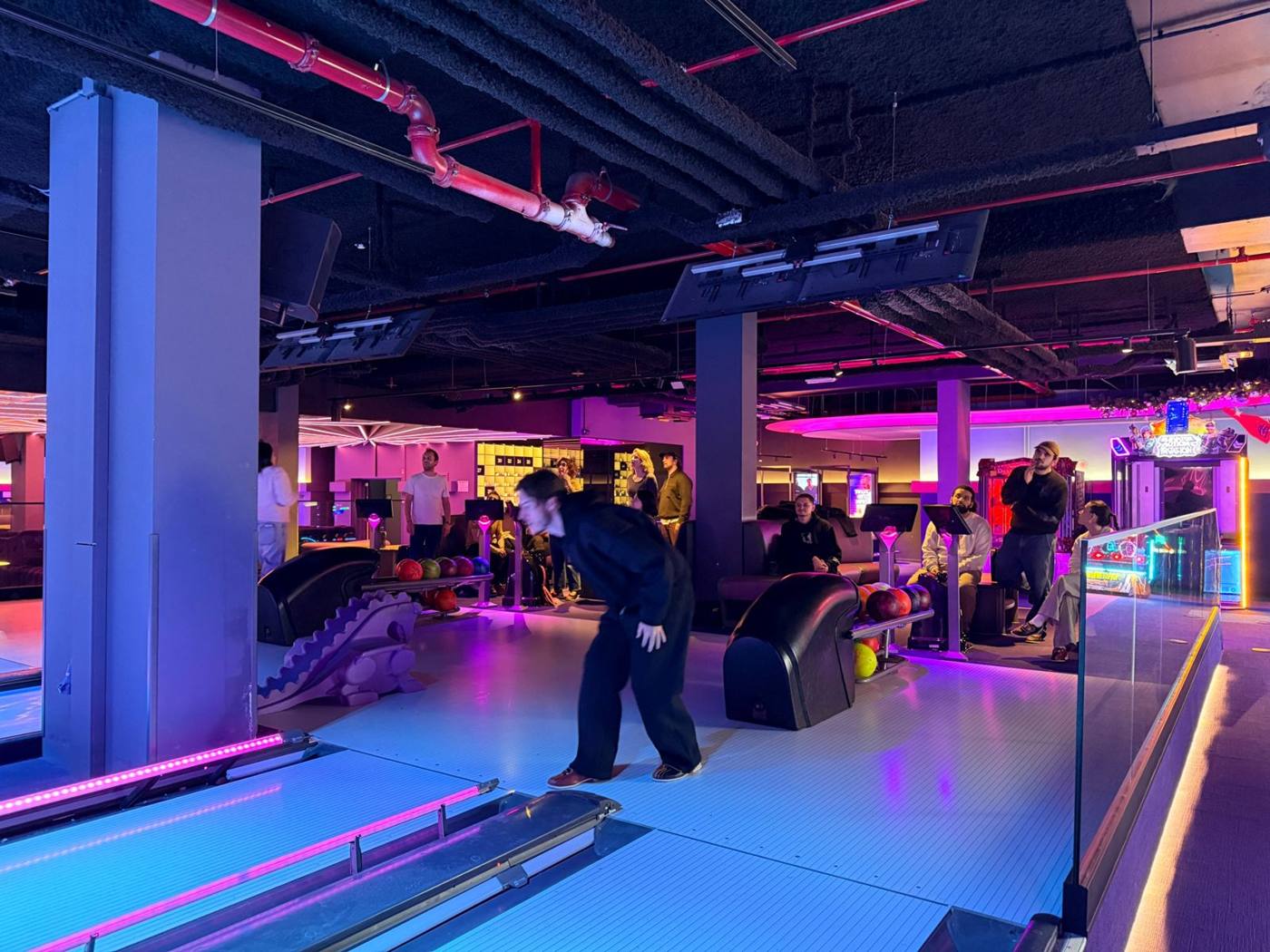 Piste de bowling éclairée en rose et violet, joueurs et écrans de score au plafond.