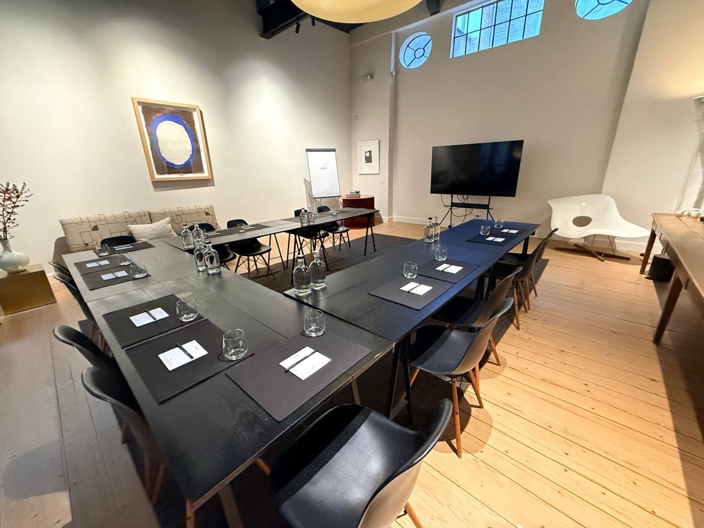 Salle de réunion en U : tables noires, carnets, verres et écran sur pied, fenêtres rondes en hauteur.
