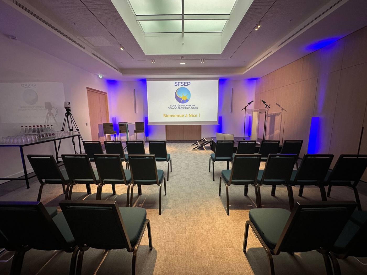 Salle de conférence à Nice : chaises en rangées, écran « Bienvenue à Nice ! », pupitre, micros et éclairage bleu.