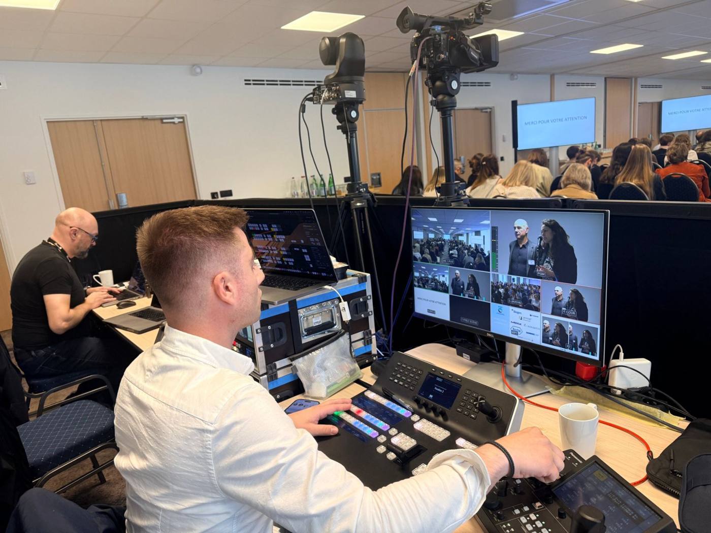 Technicien en chemise blanche au mélangeur vidéo, moniteur multi-caméras et salle de conférence pleine en arrière-plan.