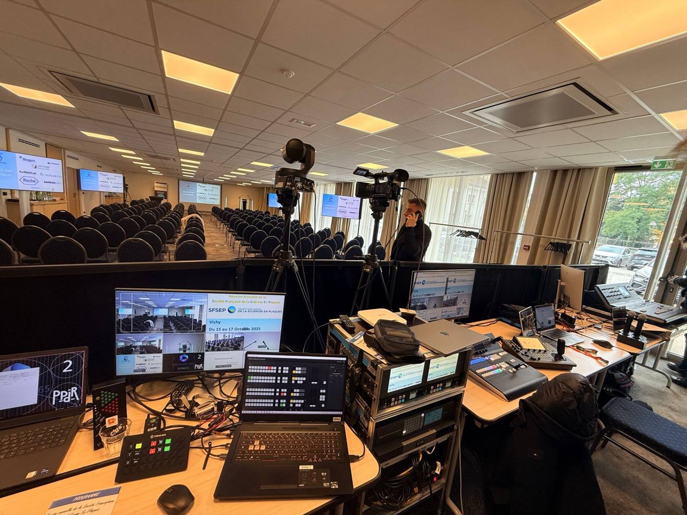Régie vidéo et son devant une salle de conférence : moniteur multi-vue, mélangeur Blackmagic ATEM, caméra motorisée et caméra d'épaule sur pied.