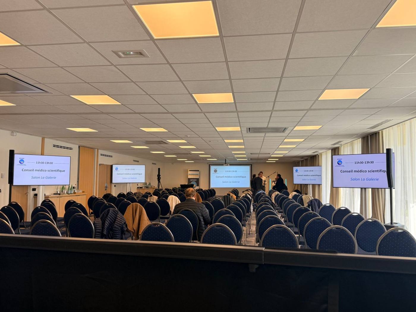 Salle de conférence « La Galerie » avec écrans de diffusion répartis dans la salle, chaises bleues et écran de projection au fond.