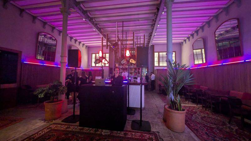DJ derrière son pupitre noir dans une halle à verrière éclairée en violet, enceintes sur pied et bar au fond.
