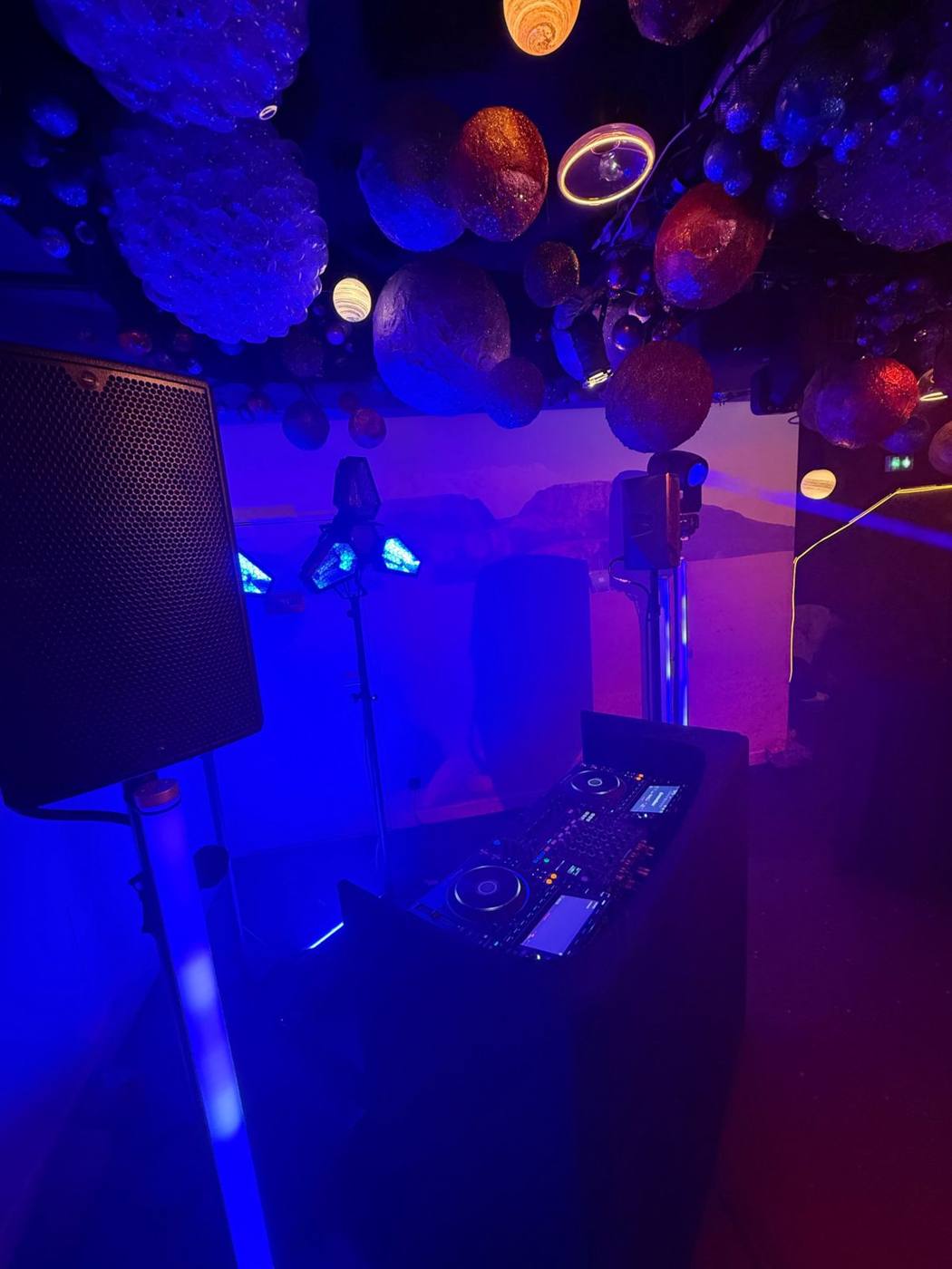 Pupitre de DJ avec contrôleur, enceinte et projecteurs sous un plafond de planètes.