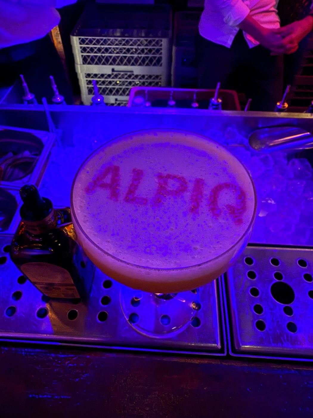 Cocktail avec le mot « ALPIQ » dessiné sur la mousse, sur un bar sous lumière bleue.