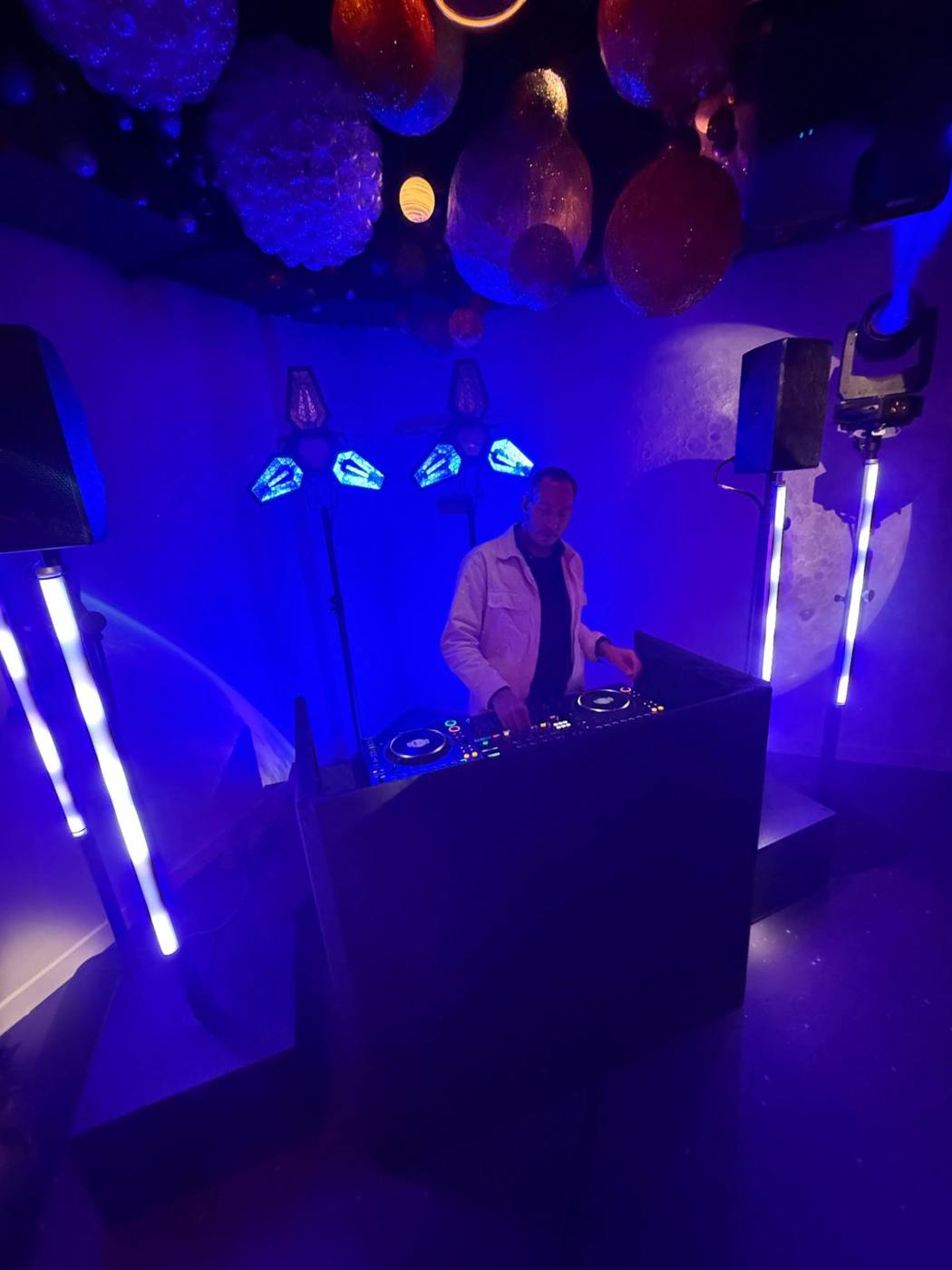 DJ derrière son pupitre, enceintes sur pied avec tubes lumineux, projecteurs et planètes au plafond.